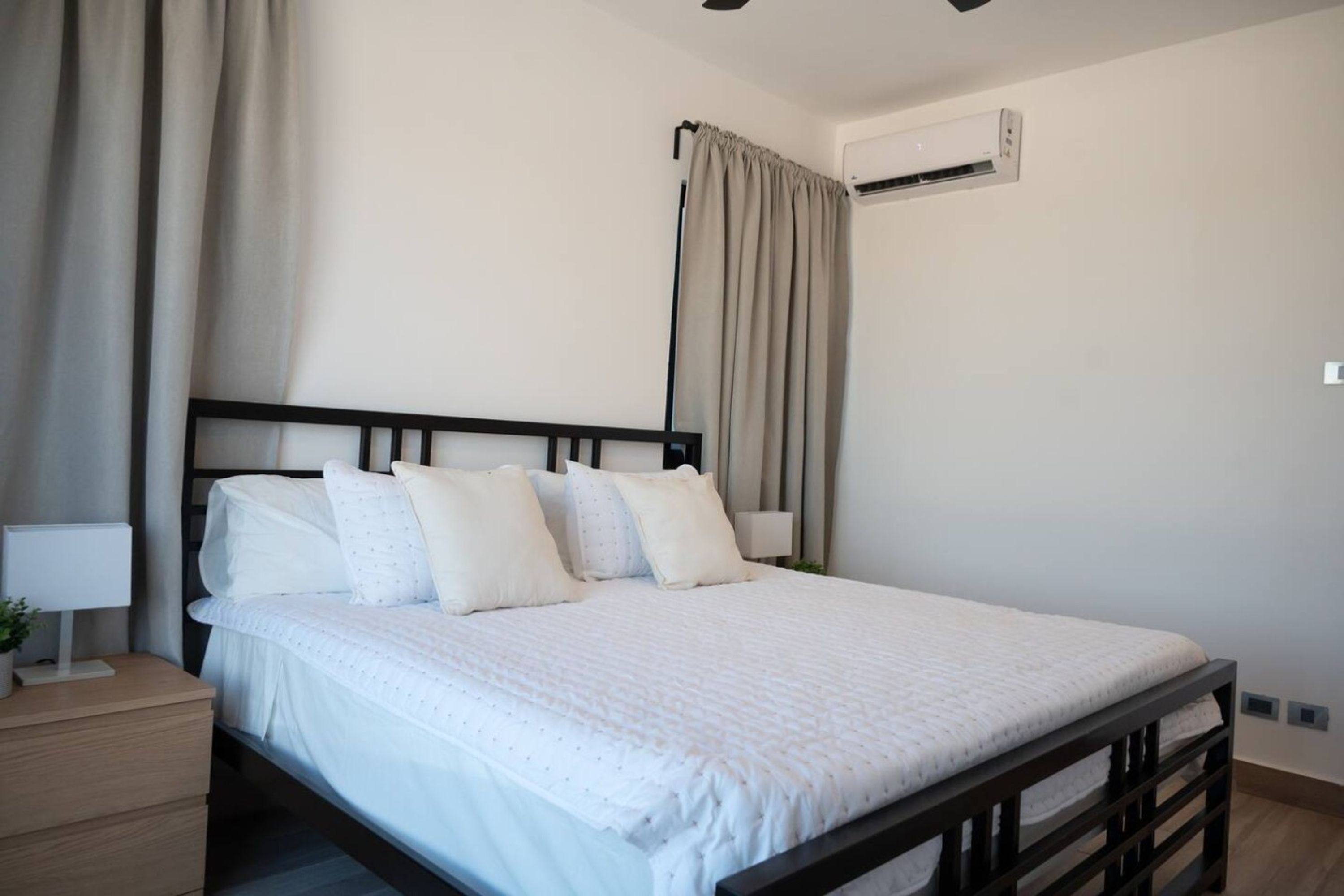 Una cama bien hecha con cabecera, una mesita de noche con lámpara y un equipo de aire acondicionado en la pared.