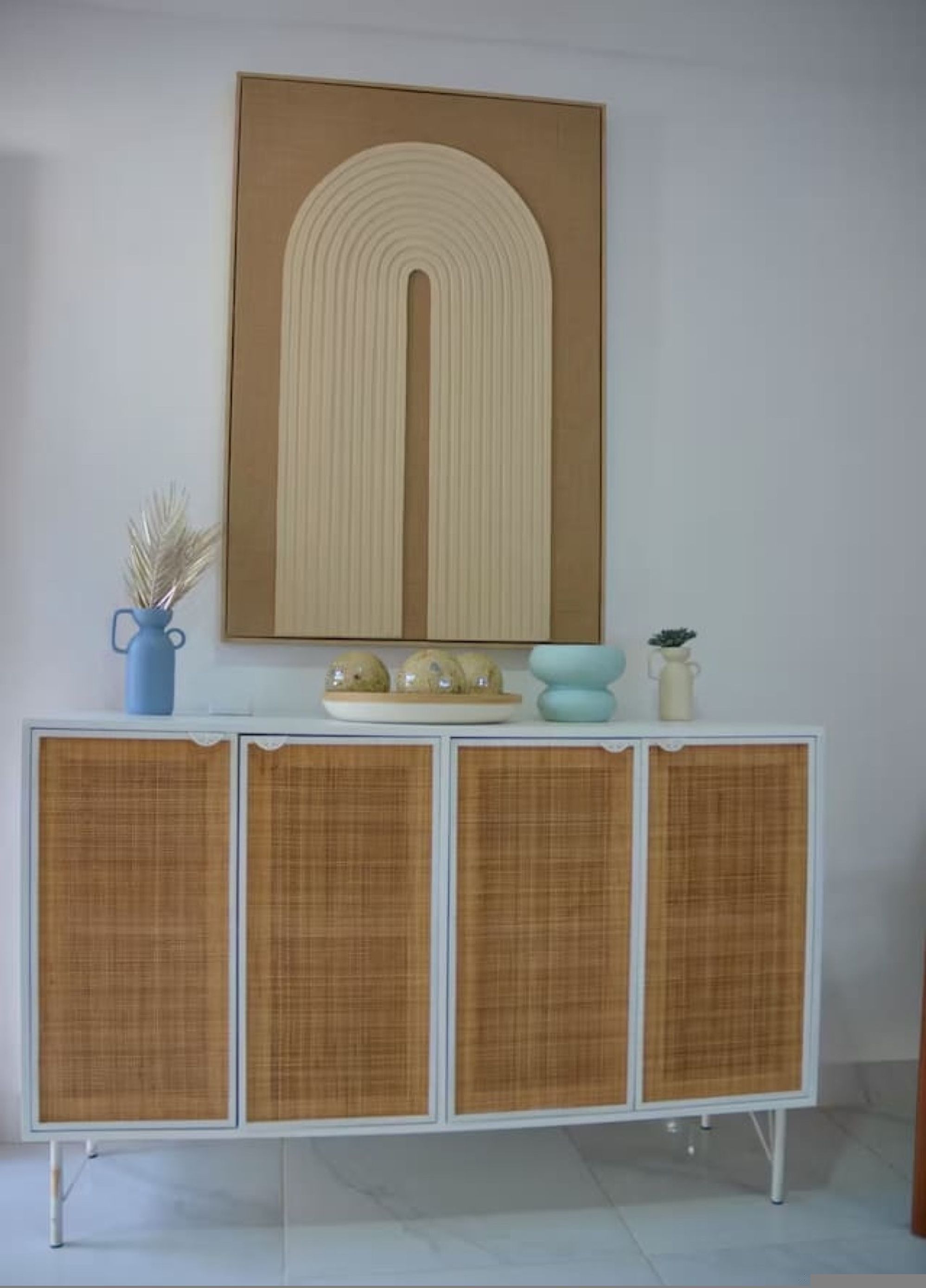 A white cabinet with four doors, each with a woven panel design. Above the cabinet, there is a large framed artwork with a vertical, curved design.