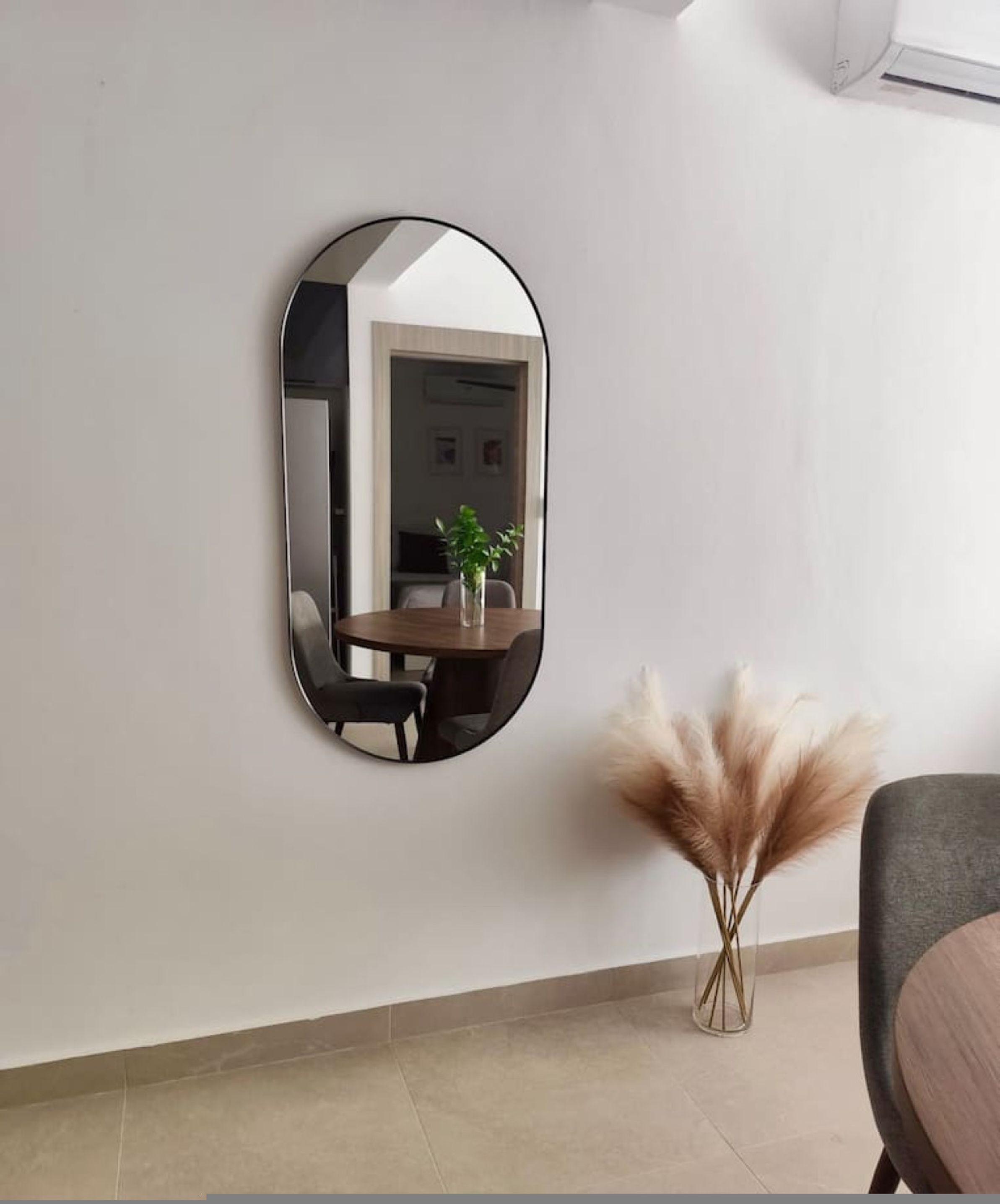 A room with a round mirror on the wall, a dining table with chairs, a vase with pampas grass, and a gray sofa.
