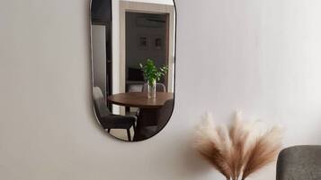 A room with a round mirror on the wall, a dining table with chairs, a vase with pampas grass, and a gray sofa.