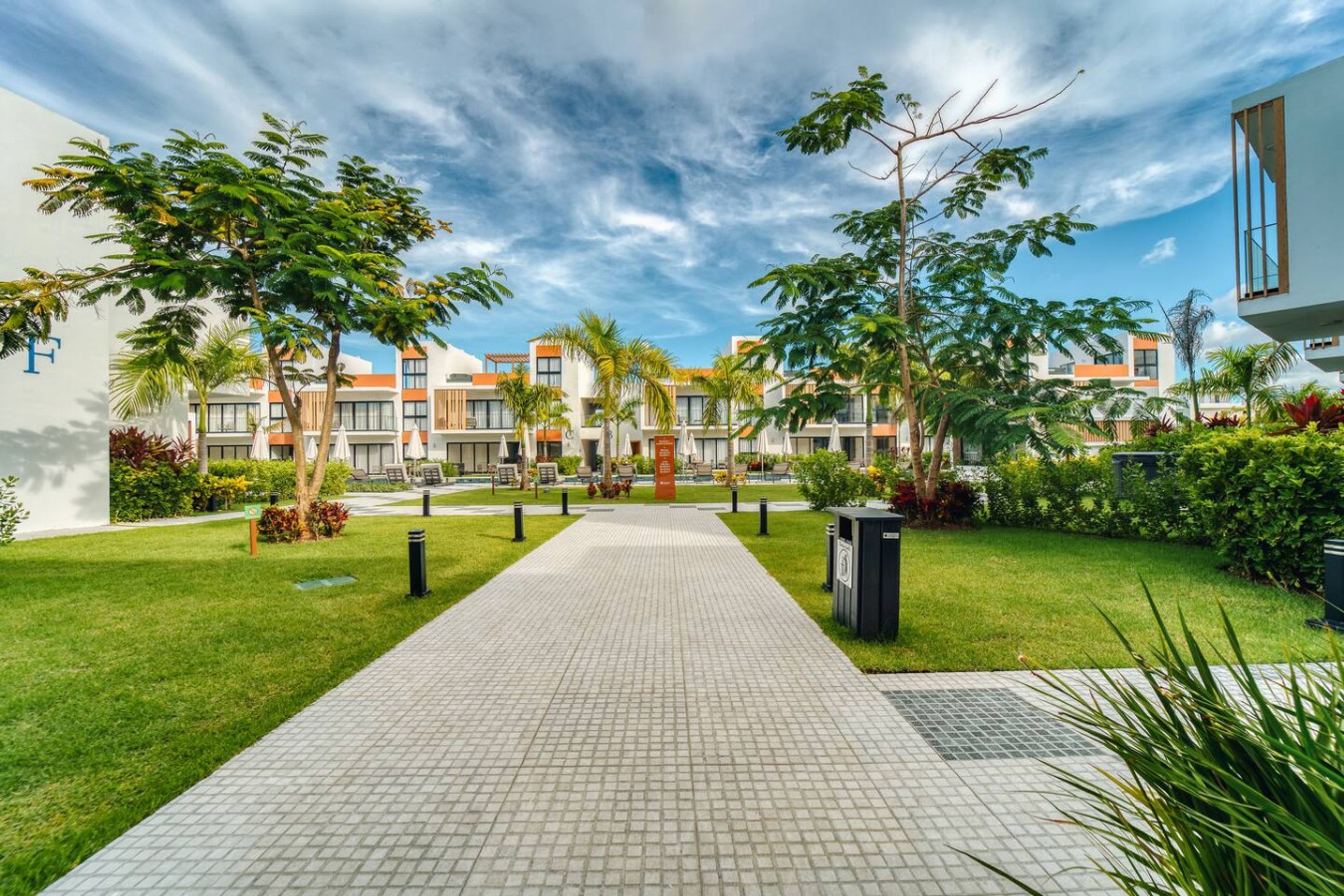 A pathway leading to a modern building with a tiled walkway, surrounded by greenery and palm trees.