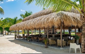 Ein Restaurant am Strand mit Reetdachschirmen, Korbmöbeln und Blick aufs Meer.