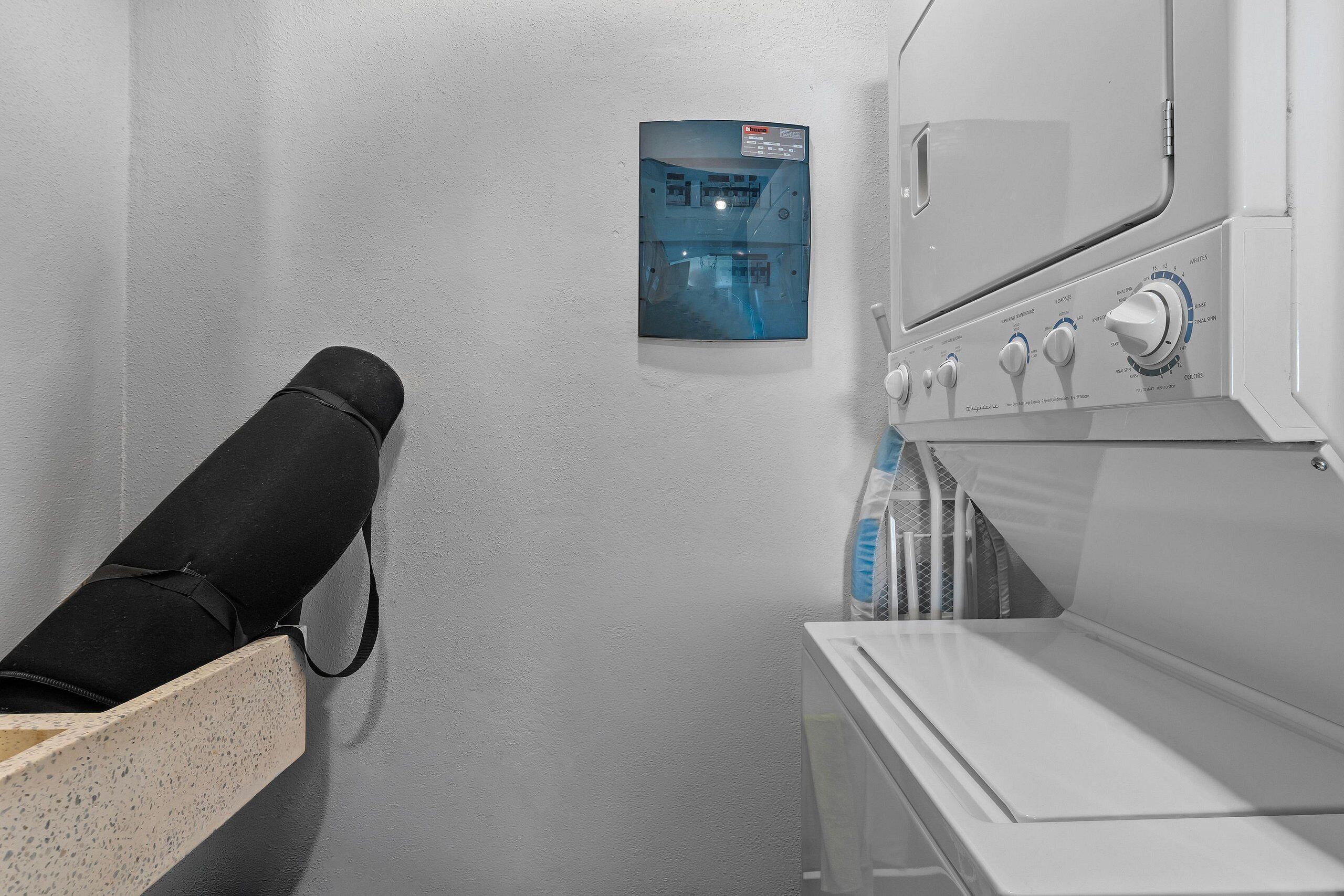 Un lavadero con un panel de control en la pared, una lavadora blanca y una maleta negra sobre un banco de madera.