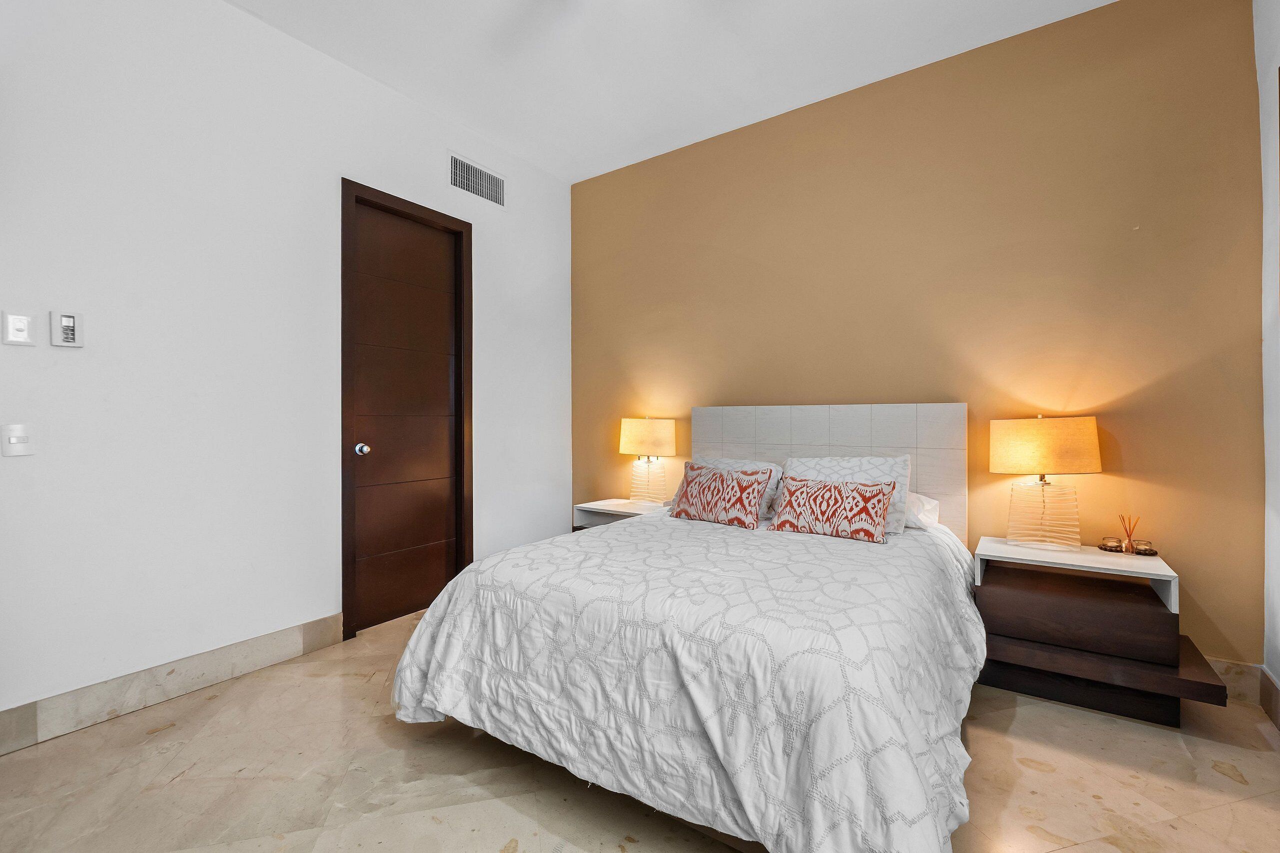 Un dormitorio con una cama, dos lámparas de noche, una mesita de noche, una puerta y un estante en la pared.