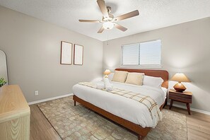 A bedroom with a bed, bedside lamps, a ceiling fan, a window with blinds, and a small table with a lamp.
