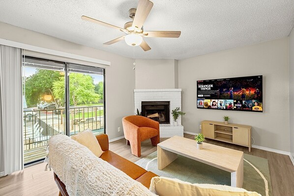 A living room with a sofa, a coffee table, a fireplace, a TV mounted on the wall, and a balcony with a view of greenery.