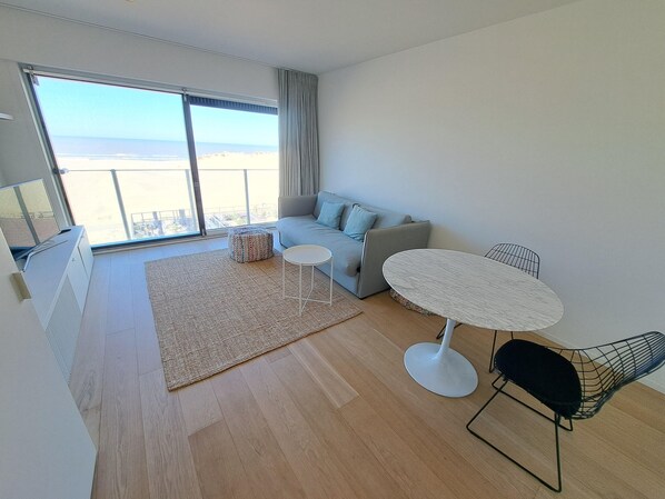 A modern living room with a sofa, a small round table, a black chair, a woven rug, and a large window offering a view of the beach. - Sea View Retreat for 2 in Oostduinkerke (Koksijde)