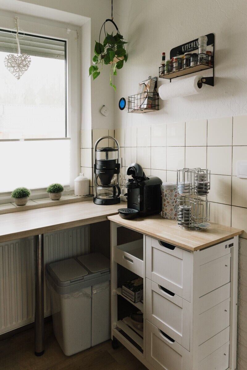 A kitchen with a coffee maker, a trash bin, a plant, and a shelf with kitchen items.