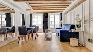 Uma sala de estar espaçosa com piso em padrão herringbone, um sofá azul, uma mesa de jantar com cadeiras e grandes janelas.