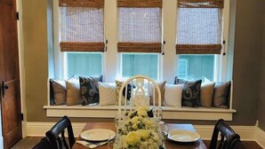Uma sala de jantar com lustre, mesa posta para seis pessoas e um assento junto à janela com almofadas.