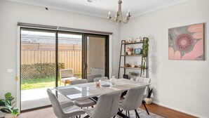 Un comedor con mesa de vidrio, sillas grises, una araña, un puertas correderas de vidrio a un patio trasero y una estantería con adornos.