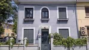 Un edificio de dos plantas con fachada gris, una entrada central con puerta y ventana, y contraventanas en las ventanas superiores.