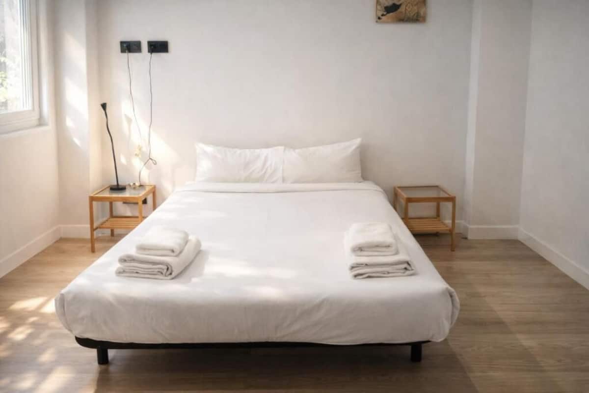 Una cama bien hecha con sábanas y toallas blancas, dos mesitas de noche, una lámpara de pie y un taburete de madera.