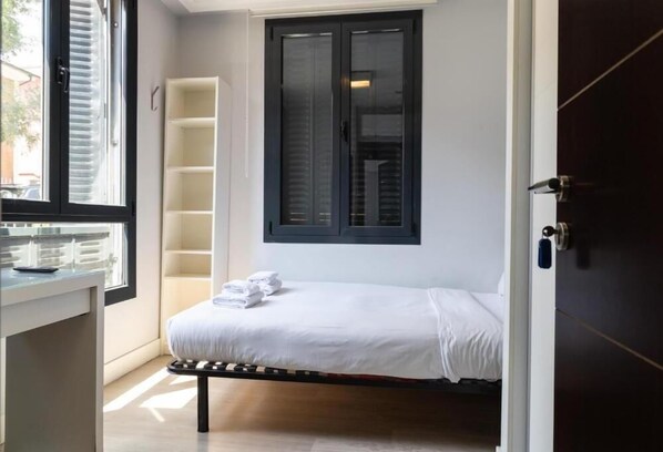 A small, single-bed room with a window, a bedside table, and a built-in shelf.