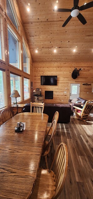 Ein Zimmer mit Holzeinrichtung, einem Esstisch, Stühlen, einem an der Wand befestigten Fernseher und einem Deckenventilator.