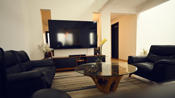 A modern living room with a large flat-screen TV, two black leather sofas, a glass coffee table, and a wooden accent piece. - ROOM WITH PRIVATE BATHROOM, IN A HOUSE WITH SHARED AMENITIES AND A POOL (Palmira)