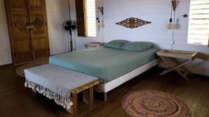 Quarto com cama, mesas de cabeceira, um banquinho, um ventilador e um objeto decorativo na parede.