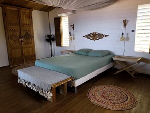 A bedroom with a bed, bedside tables, a bench, a fan, and a decorative wall piece. - Ayana Lodge - Sea view &clim Bouillante (Bouillante)