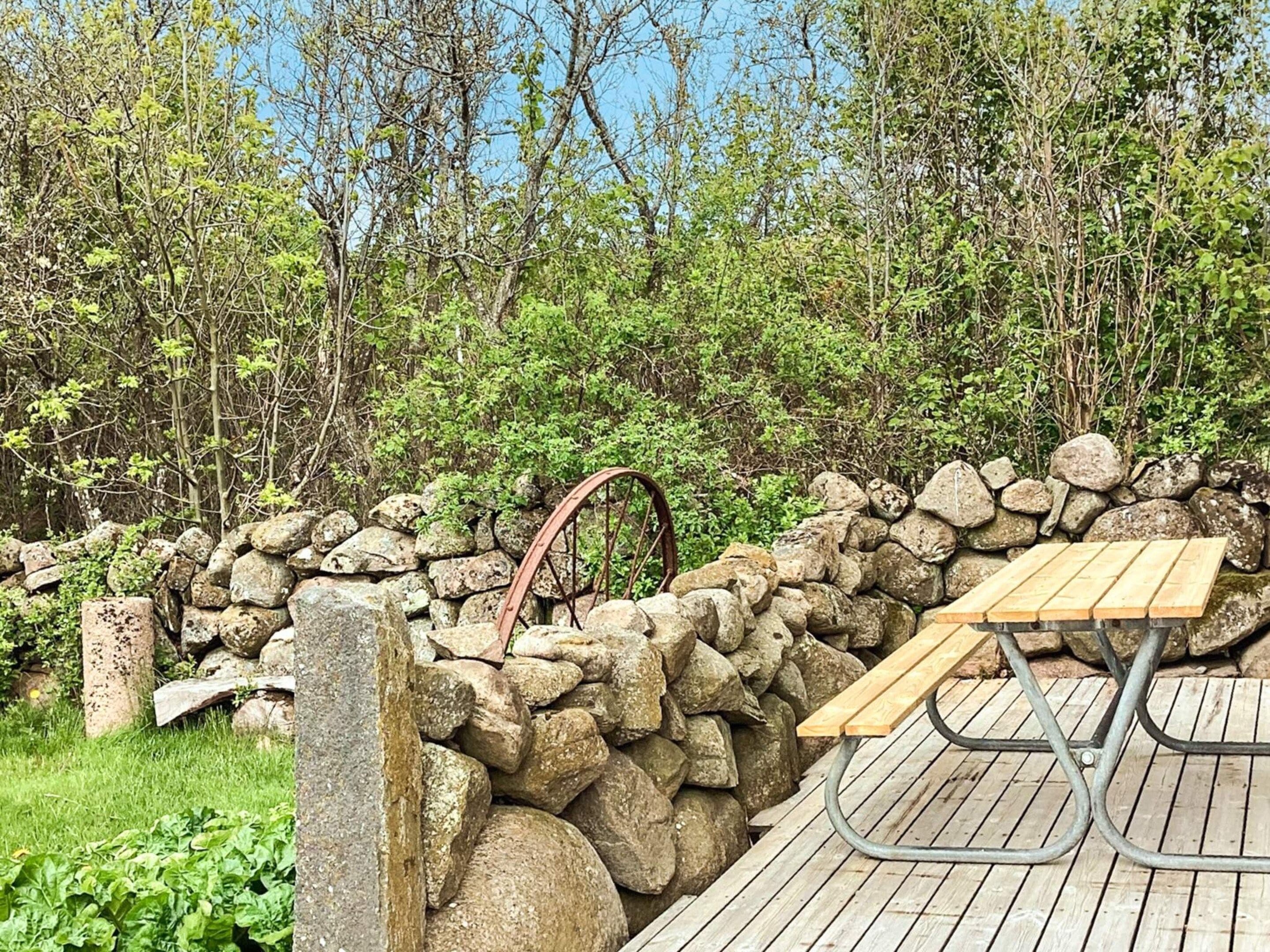 A stone wall with a wooden bench and a metal table, surrounded by greenery and trees.