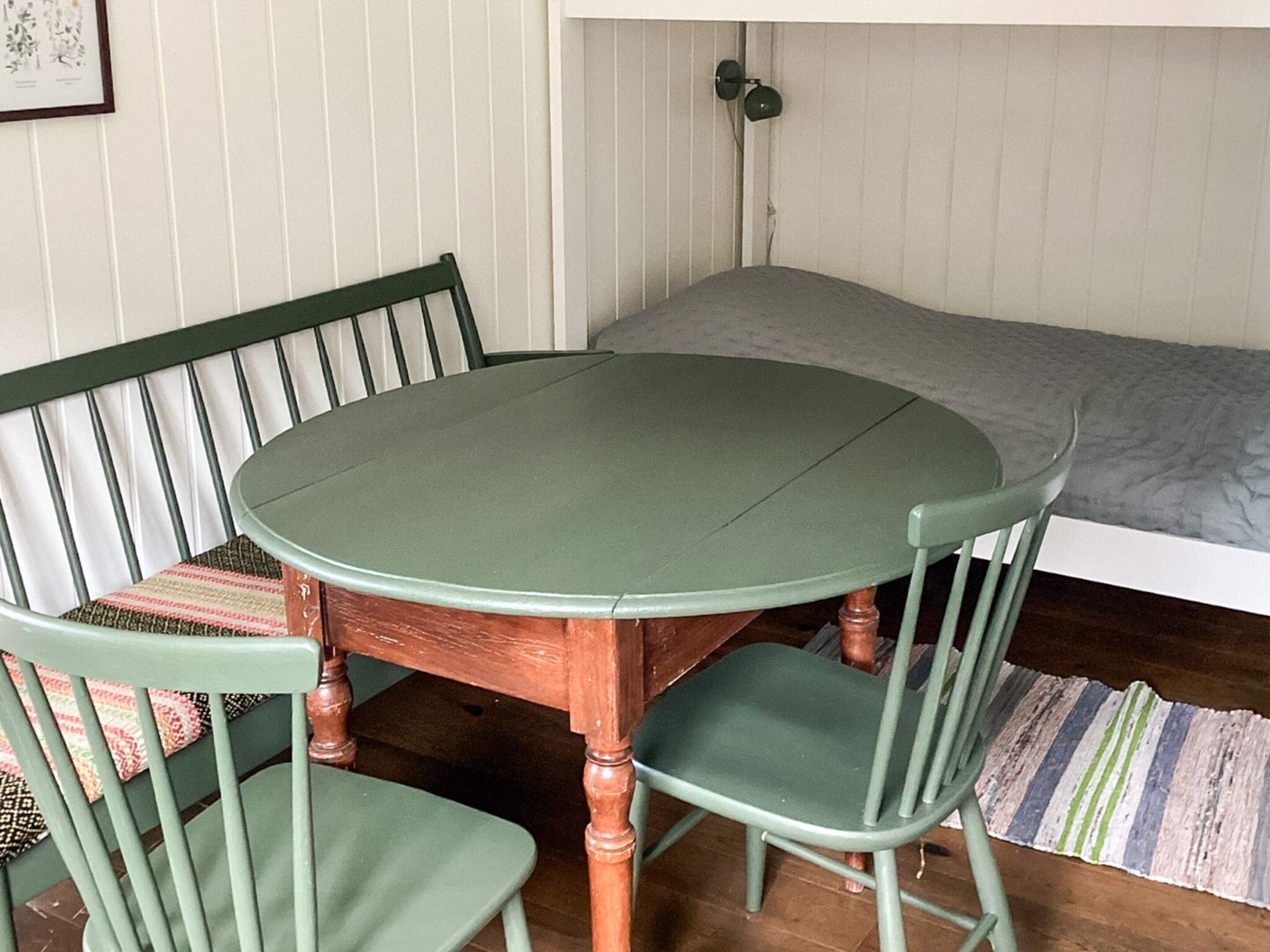 Une pièce avec une table ronde en bois, deux chaises vertes, un lit avec un matelas gris et un tapis à rayures.