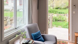 Una habitación con un sillón gris, una mesita con un control remoto y vistas a un jardín por la ventana.