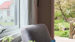 Una habitación con ventana, una silla gris, un almohá de color azul y vista a un jardín.