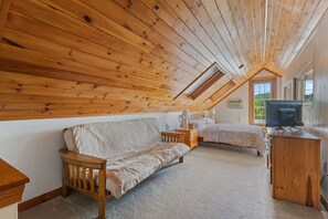 Ein gemütliches Schlafzimmer im Dachgeschoss mit schrägen Wänden, einer Schlafcouch, einem Bett und einem Fernseher auf einer Holzkommode.