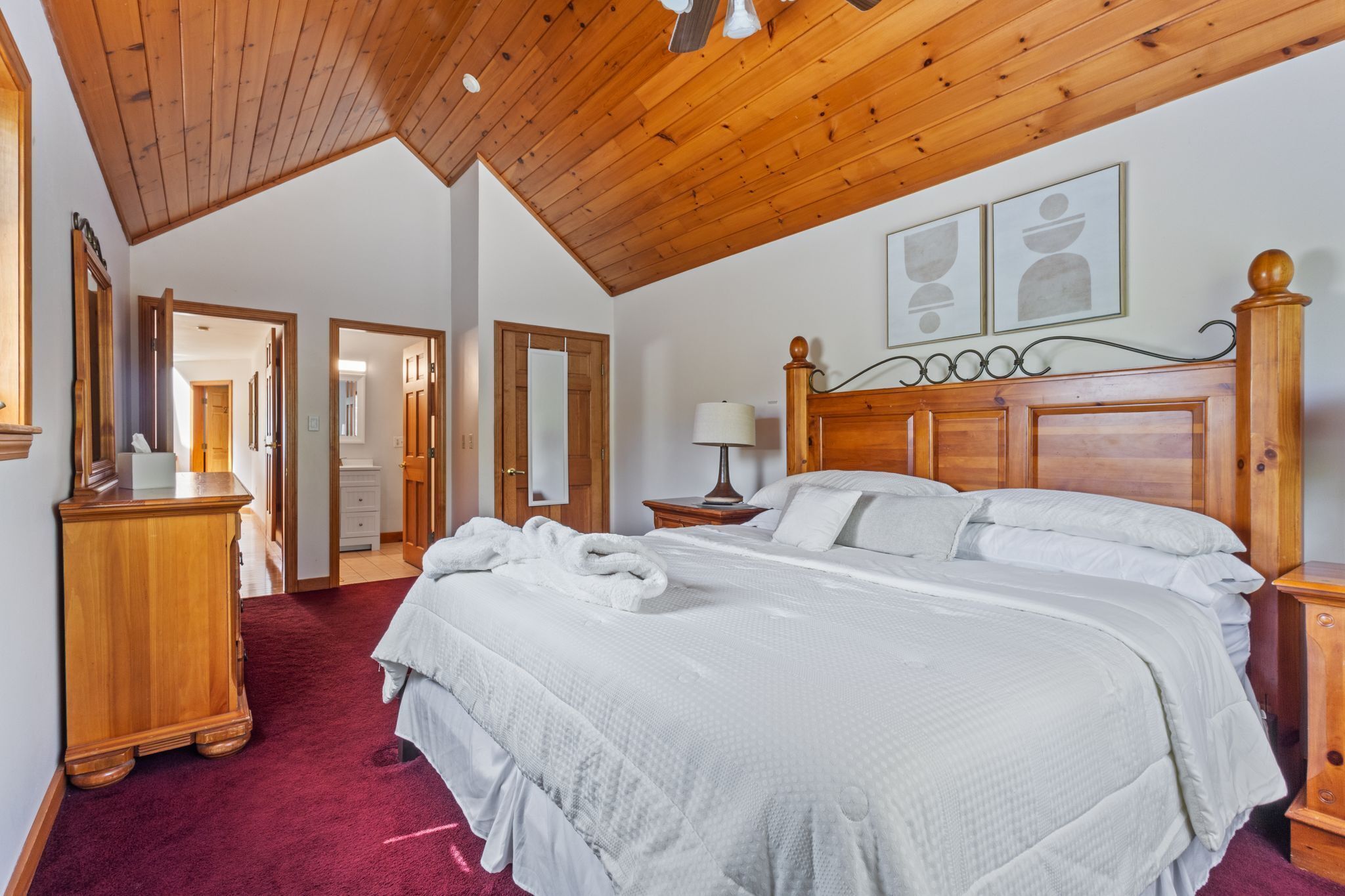 Quarto com teto de madeira, uma cama grande com roupa de cama branca, uma mesa de cabeceira de madeira e um espelho na parede.