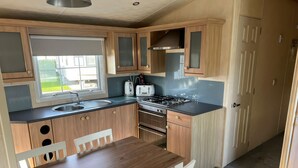 A modern kitchen with wooden cabinets, a sink, a stove, and a dining area with a table and chairs. - 261 @ Owens Caravan Park (Abergele)