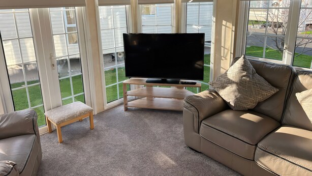 A living room with a television, a sofa, a bench, and large windows. - 261 @ Owens Caravan Park (Abergele)