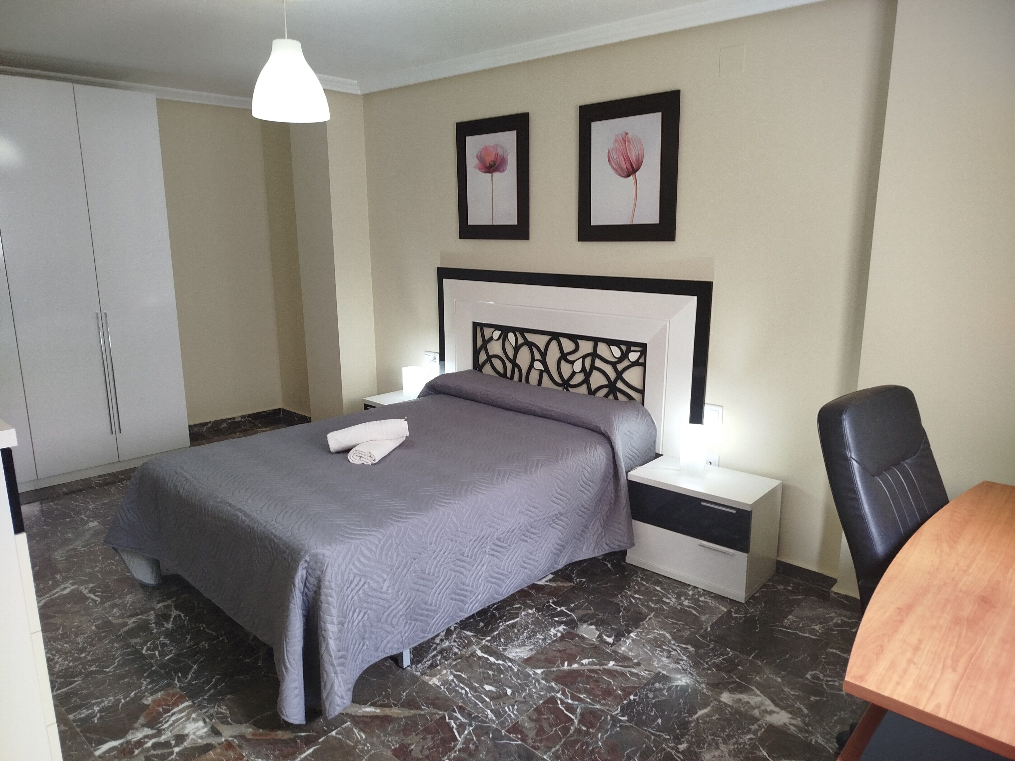 Ein Hotelzimmer mit Bett, Nachttischen, Schreibtisch, Stuhl, Kleiderschrank und zwei gerahmten Bildern an der Wand.