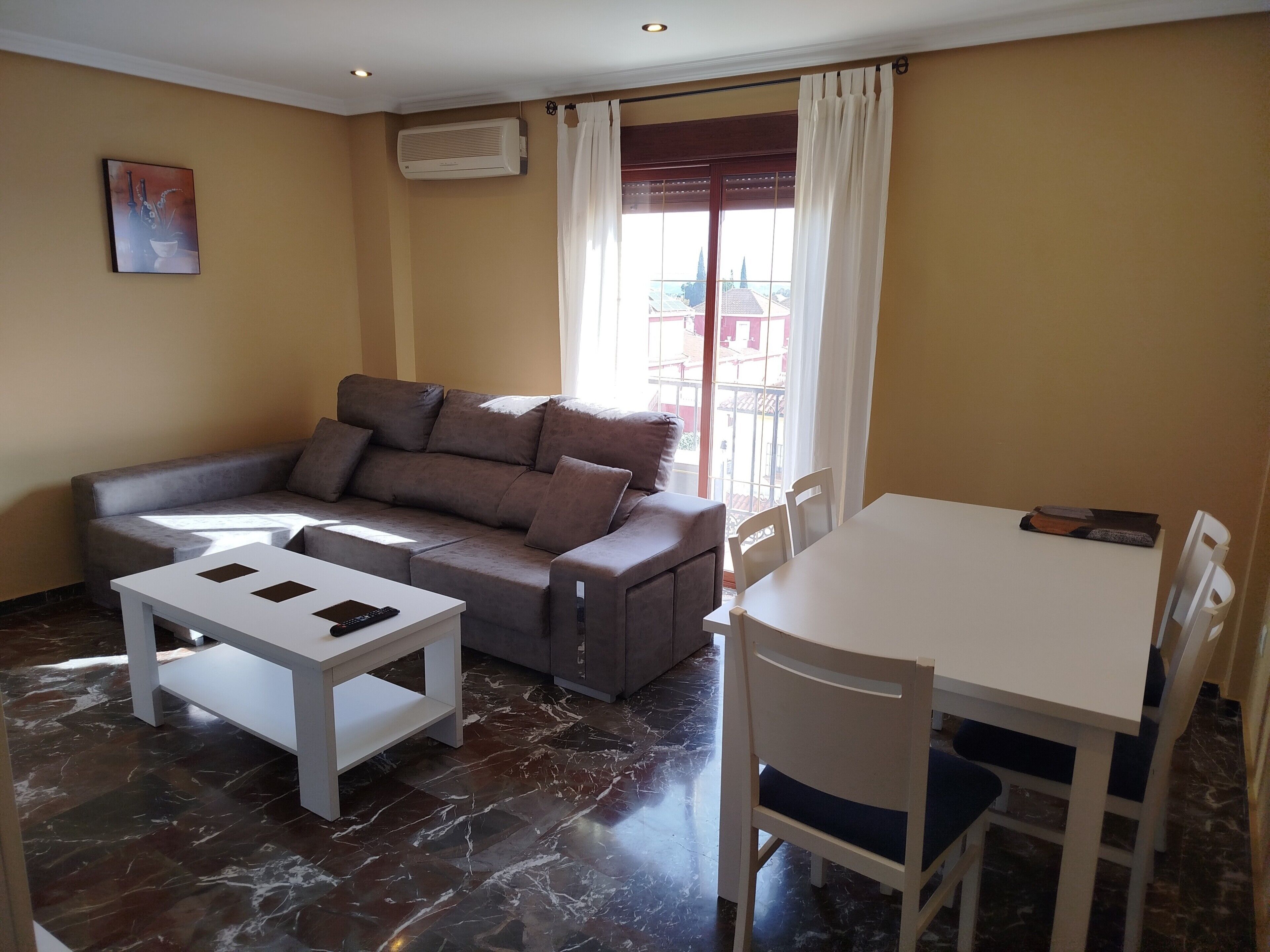 A living room with a sofa, a small table, a dining table with chairs, a painting on the wall, and an air conditioning unit.