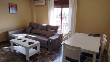 A living room with a sofa, a small table, a dining table with chairs, a painting on the wall, and an air conditioning unit.