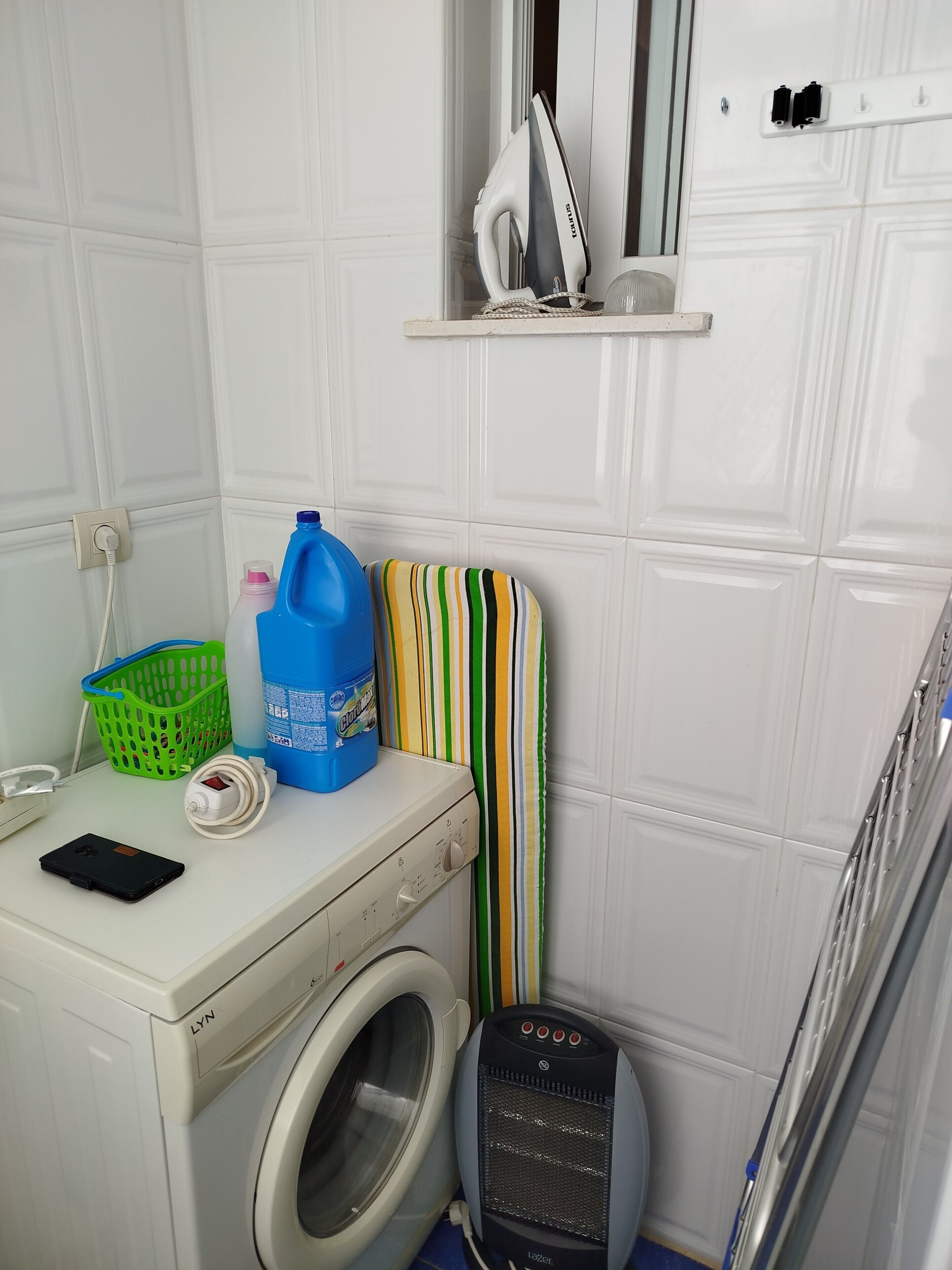 Un espace dédié à la lessive, comprenant un lave-linge, un sèche-linge, un panier à linge vert, une bouteille de lessive bleue et une planche à repasser à rayures.