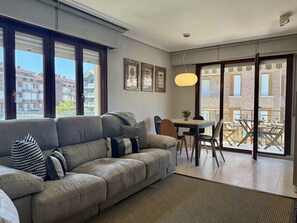 A modern living room with a sofa, a dining table, and a view through large windows. - Apartment Bajosaltos 6905 (Noja)