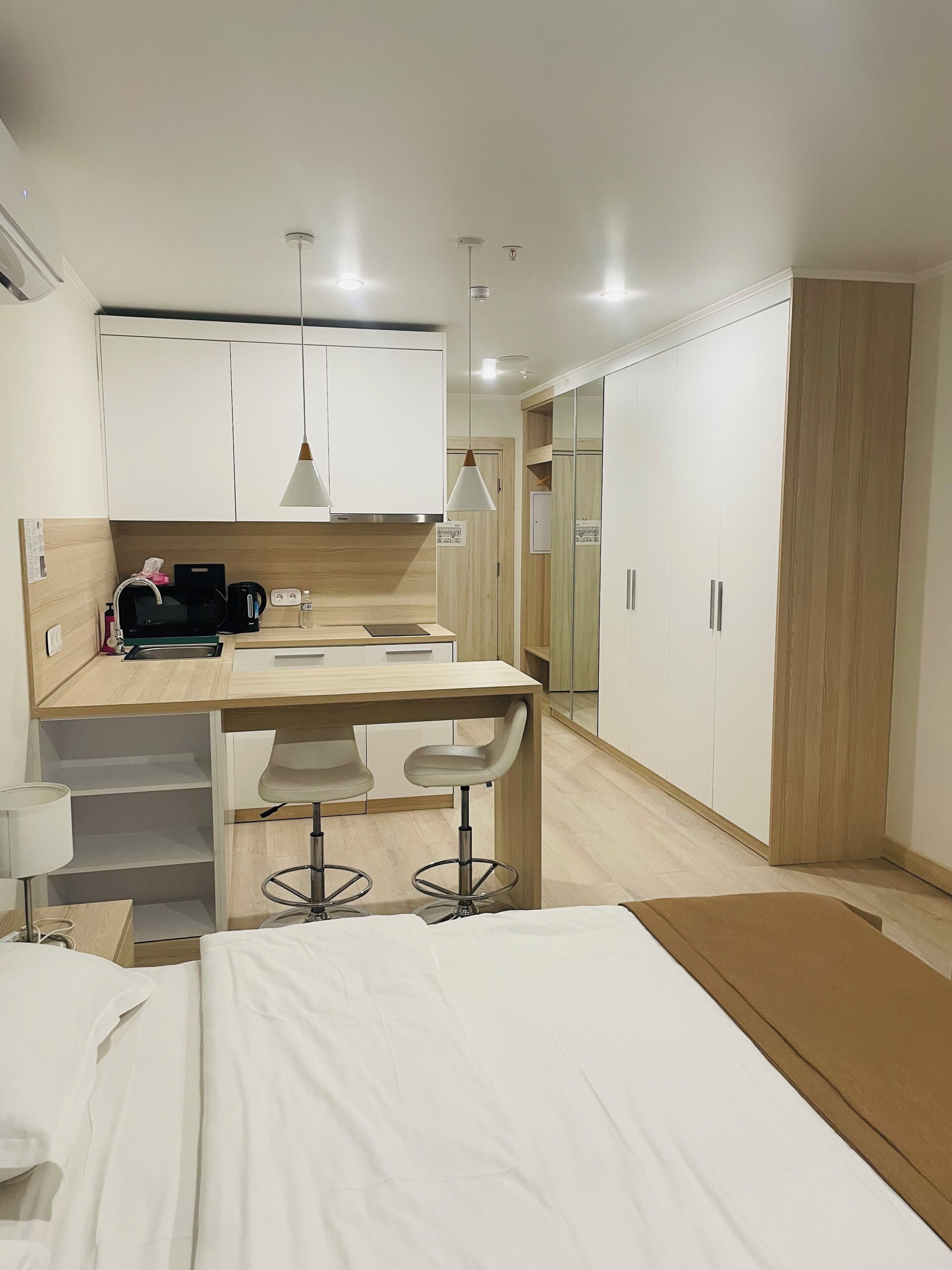 A modern hotel room with a bed, a desk with a chair, a kitchenette, and a wardrobe.