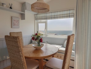 Un comedor con una mesa redonda de madera, sillas de mimbre, un jarrón con flores, un ventanal con vistas al mar y un cartel con la inscripción 'BON APPETIT' en la pared.