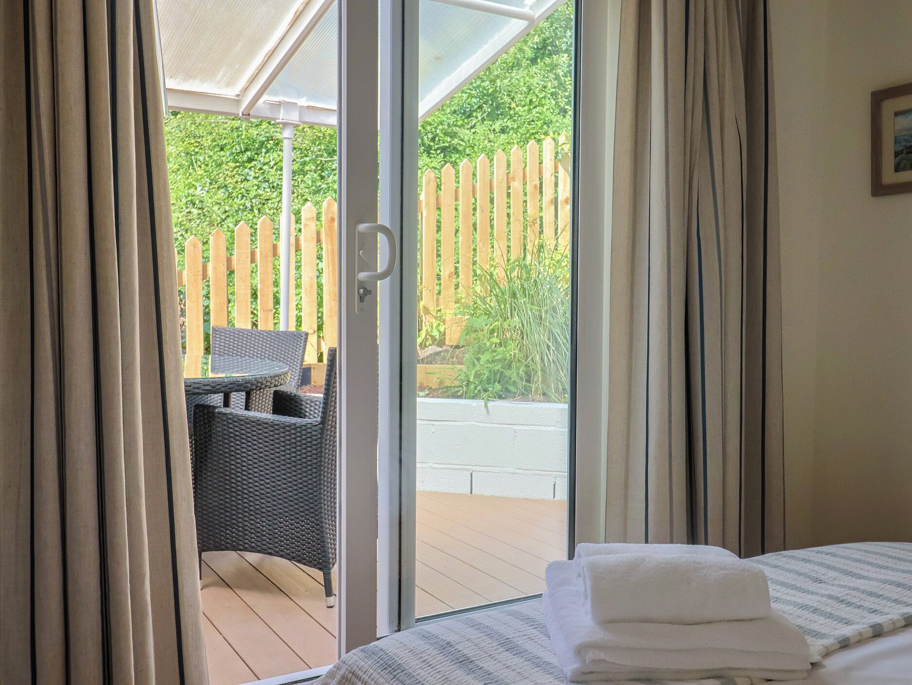 Quarto com cama, janela com cortinas e vista para uma cerca de madeira e vegetação lá fora.