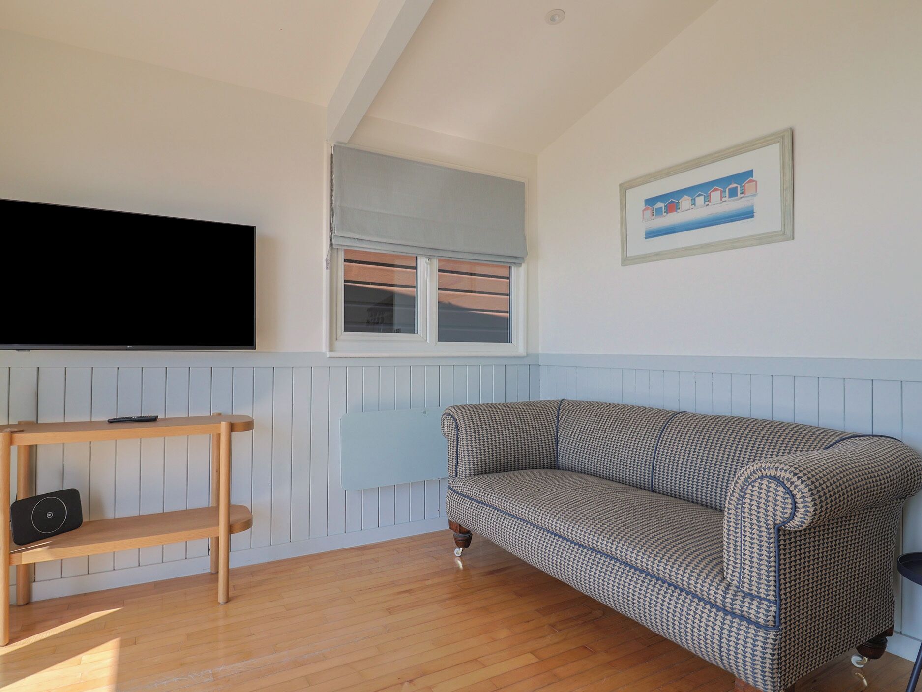 Sala de estar com sofá, televisão na parede, janela com persianas, quadro emoldurado e prateleira de madeira.
