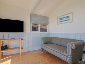 Sala de estar com sofá, televisão na parede, janela com persianas, quadro emoldurado e prateleira de madeira.