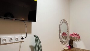 Una habitación con escritorio de madera, televisor montado, aire acondicionado, espejo y toma de corriente con cable.
