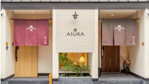 Ein Hotel Eingang mit einem Schild mit der Aufschrift 'ASUKA' und zwei Türen, eine mit einer Holzfassade und die andere mit einer beige farbenen Fassade.