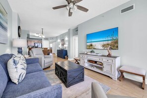 A living room with a sofa, coffee table, and TV. There is a kitchen in the background. - Sunrise Beach #1203 (Panama City Beach)