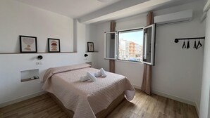 Un dormitorio con una cama, un ventanal con vistas a edificios, un perchero y una estantería con un objeto pequeño.
