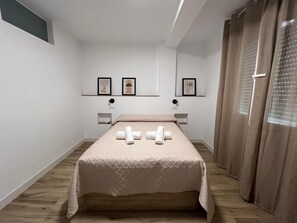 Un dormitorio con una cama, dos mesitas de noche, una lámpara de pared, un ventanal con cortinas y dos cuadros en la pared.