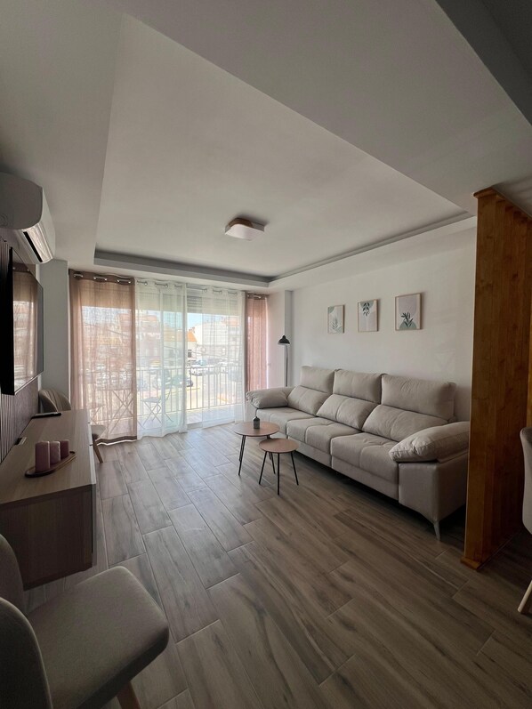 Una sala de estar con sofá, mesa de centro, televisor y un amplio ventanal con cortinas.