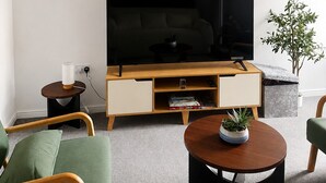 Uma sala de estar moderna com uma televisão de tela plana, um móvel de entretenimento de madeira, poltronas verdes, uma mesinha lateral e um relógio fixado na parede.