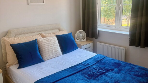 Quarto com cama, mesa de cabeceira, ventilador, janela com cortinas e um quadro pendurado na parede.