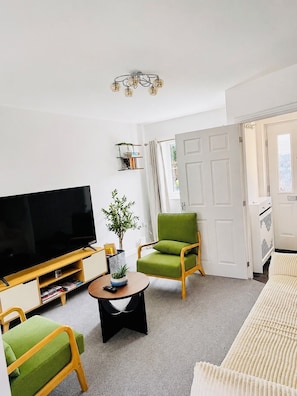 A living room with a flat-screen TV, green armchairs, a coffee table, and a bookshelf.