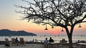 Un espacio de estar frente a la playa con sillas y mesas, un árbol y vista al mar con barcos.